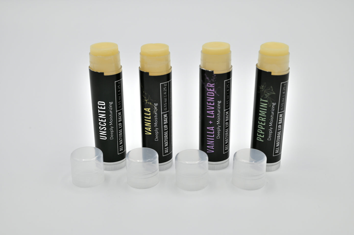 4 all natural lip balms with black labels standing upright in a line open showing a clear balm and clear caps sitting at each base on a white background