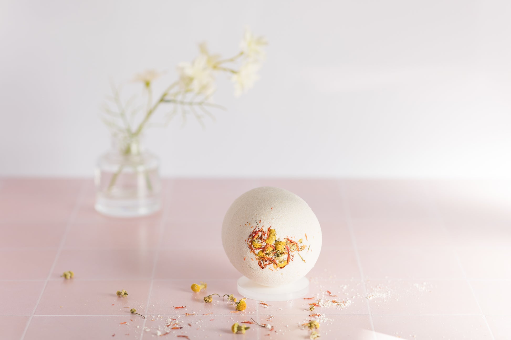 bath bomb on tile with dried chamomile buds and safflower petals scattered around it with a jar and a flower in the background