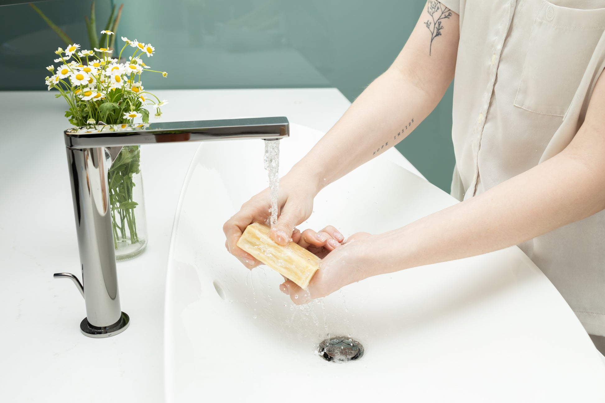 square white and yellow swirled soap being held by two hands under the faucet with running water in the sink by a bouquet of chamomile flowers
