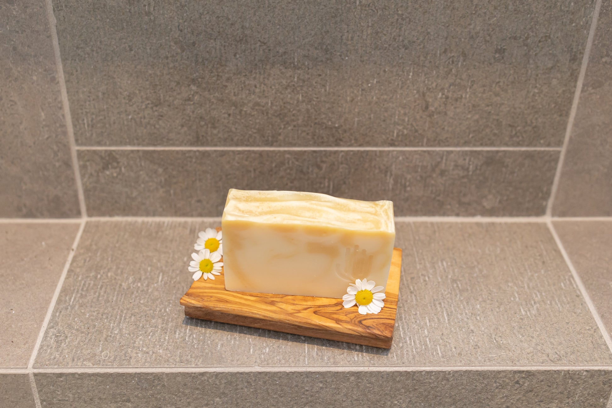 square white and yellow swirled soap placed on a wooden soap dish on shower tiles with chamomile flowers laying on the dish
