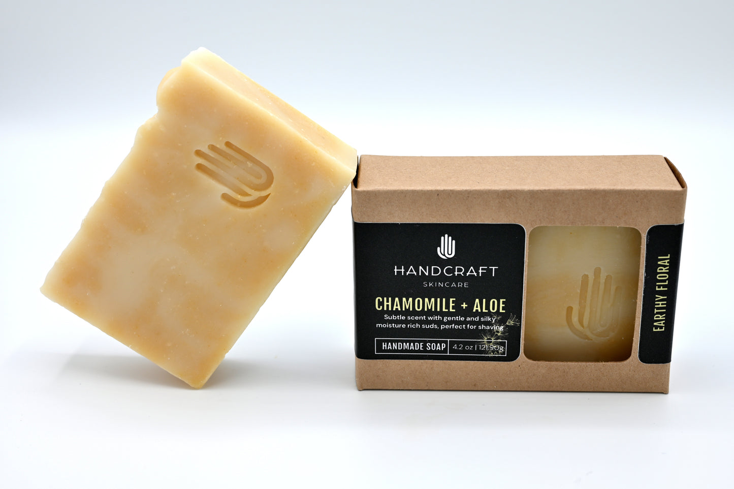 yellow and white swirled chamomile and aloe soap is unpackaged and leaning at an angle on a packaged chamomile and aloe soap in a Kraft box with a black label