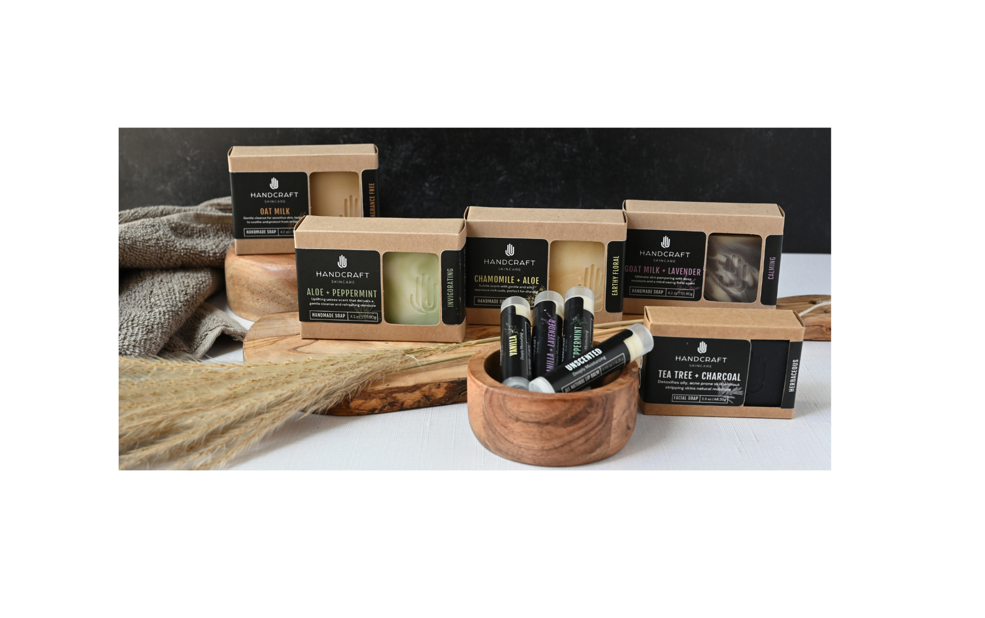 variety of 5 soaps packaged in Kraft boxes with black labels sitting on a wooden cutting board with 4 lip balms sitting in a wooden bowl in front