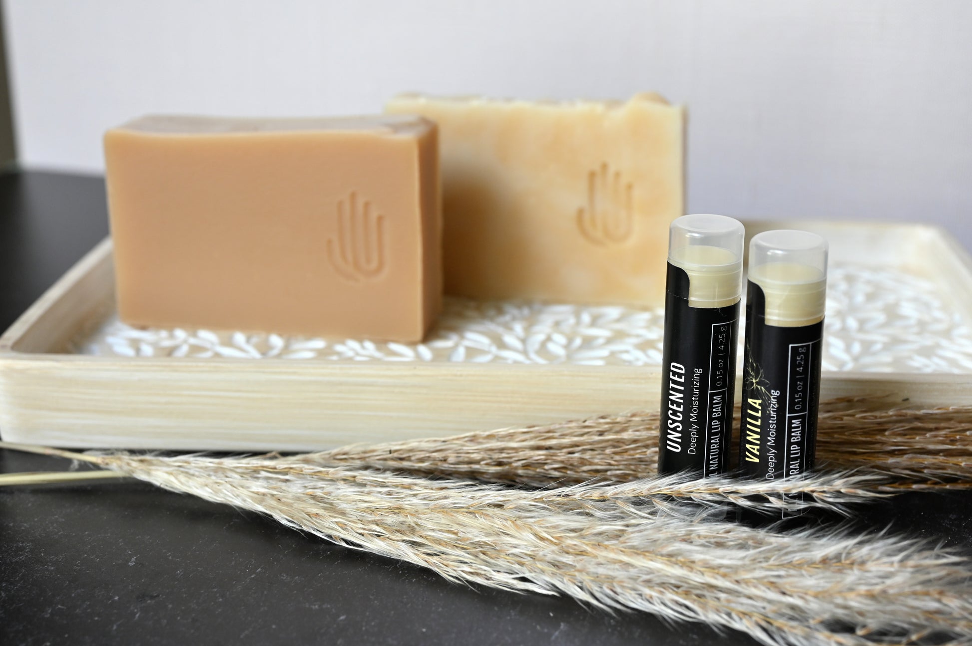 2 all natural lip balms with black labels sitting upright in dried decorative grass stalks in front of a decorative wooden tray with 2 tan soaps sitting on it