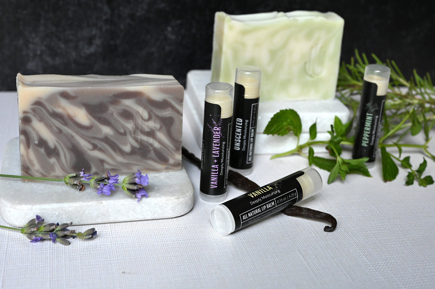 4 lip balms on a white riser surrounded by fresh herbs and matching green and white peppermint and white purple and lavender goat milk and lavender soaps