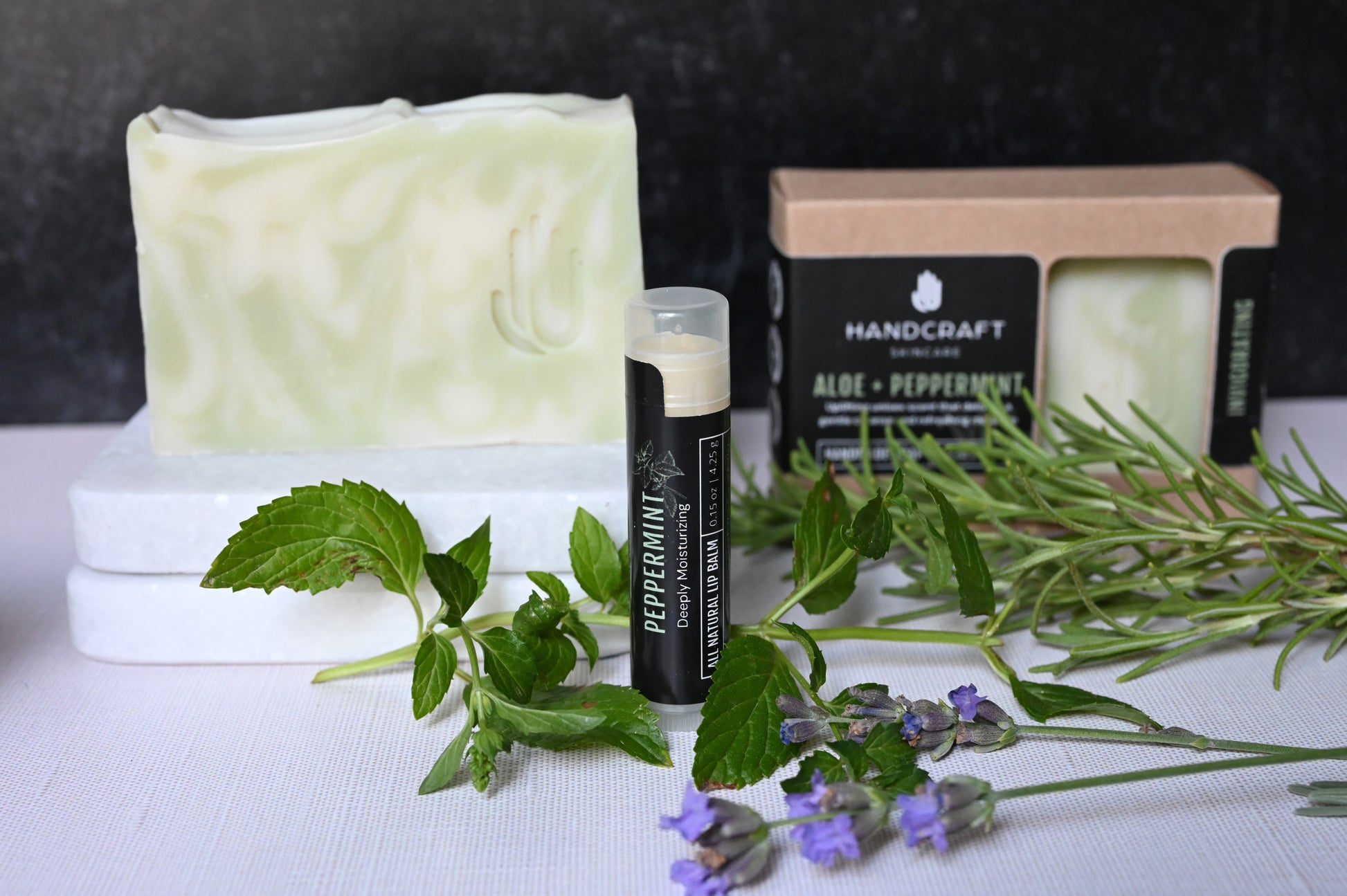 All natural peppermint lip balm standing upright with a black label surrounded by fresh rosemary and peppermint herbs and a matching peppermint bar soap