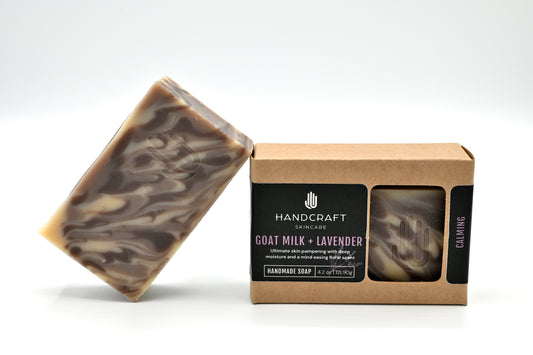 purple lavender and white swirled goat milk and lavender soap is unpackaged and leaning at an angle on a packaged goat milk and lavender soap in a Kraft box with a black label