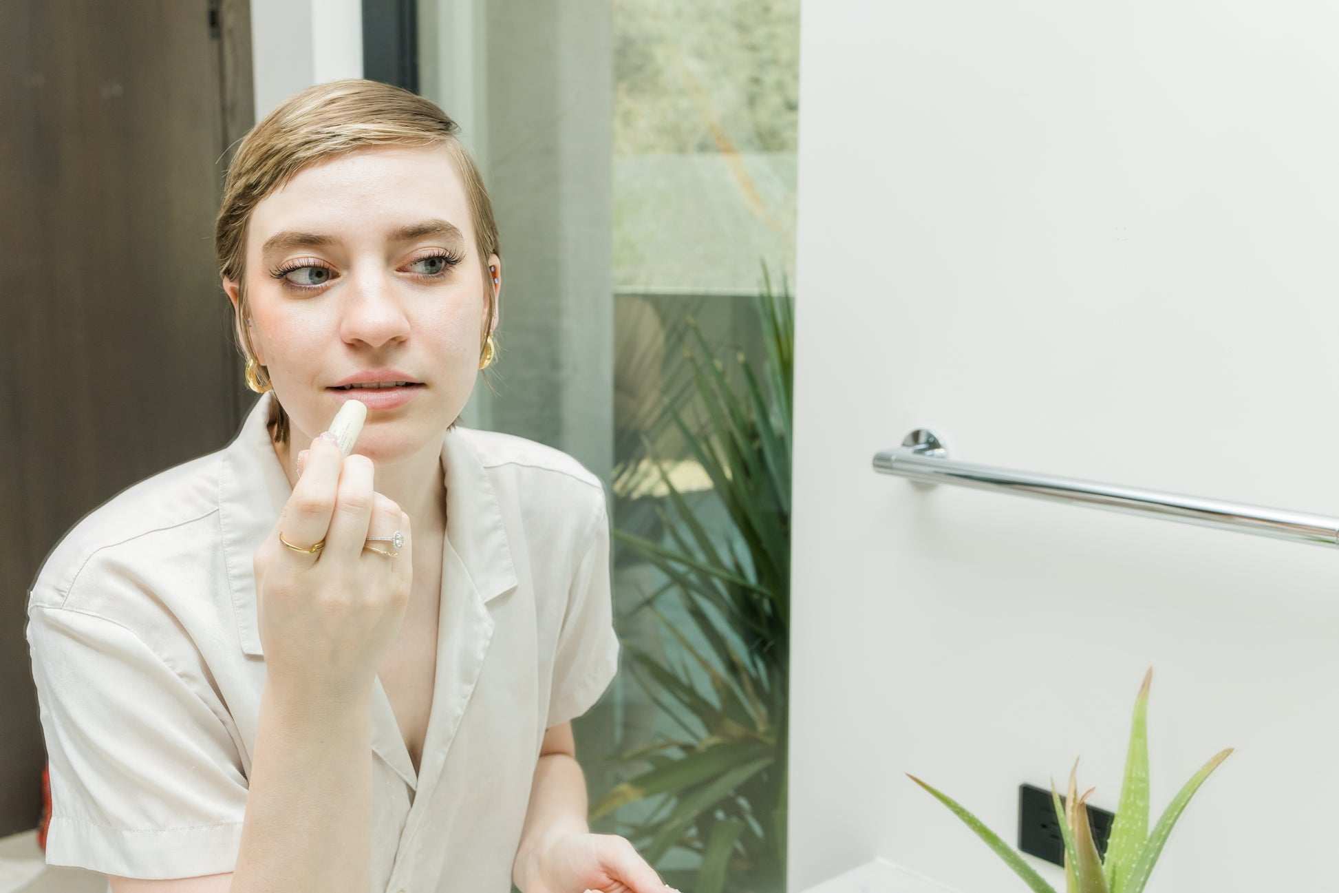 woman applying lip balm in the mirror with plants in the background