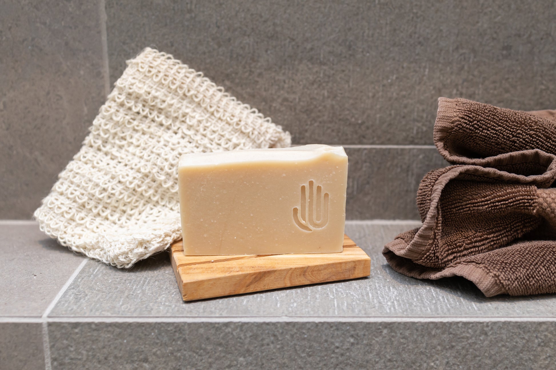 square tan colored soap on a wooden soap dish placed on shower tile with tan and brown towels in the background