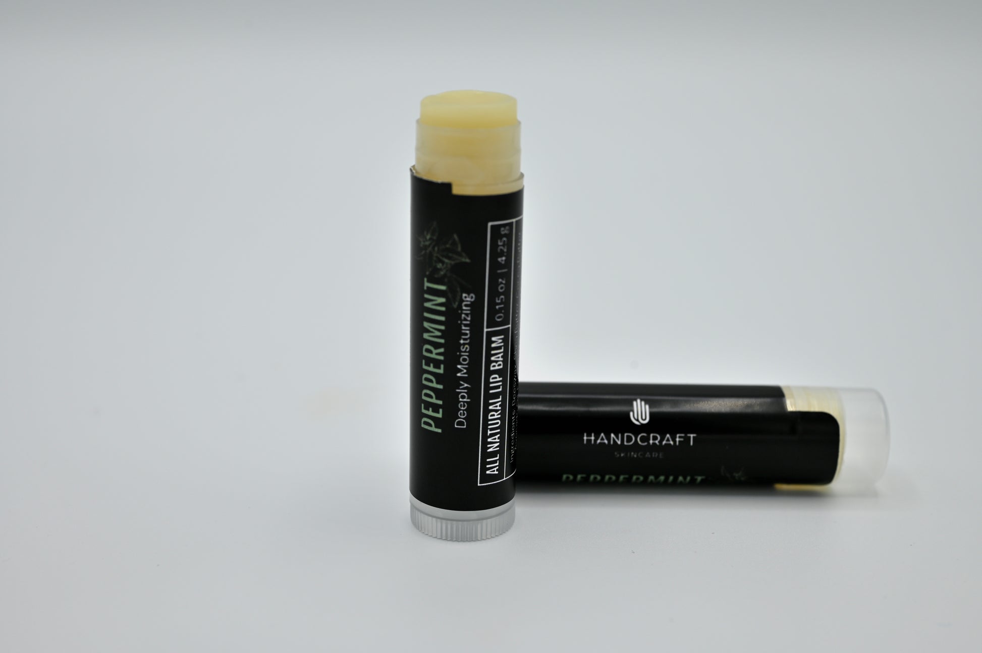 2 all natural peppermint lip balms with a black labels with one open showing a clear balm and sitting upright and one is sitting on its side closed behind the open tube of lip balm