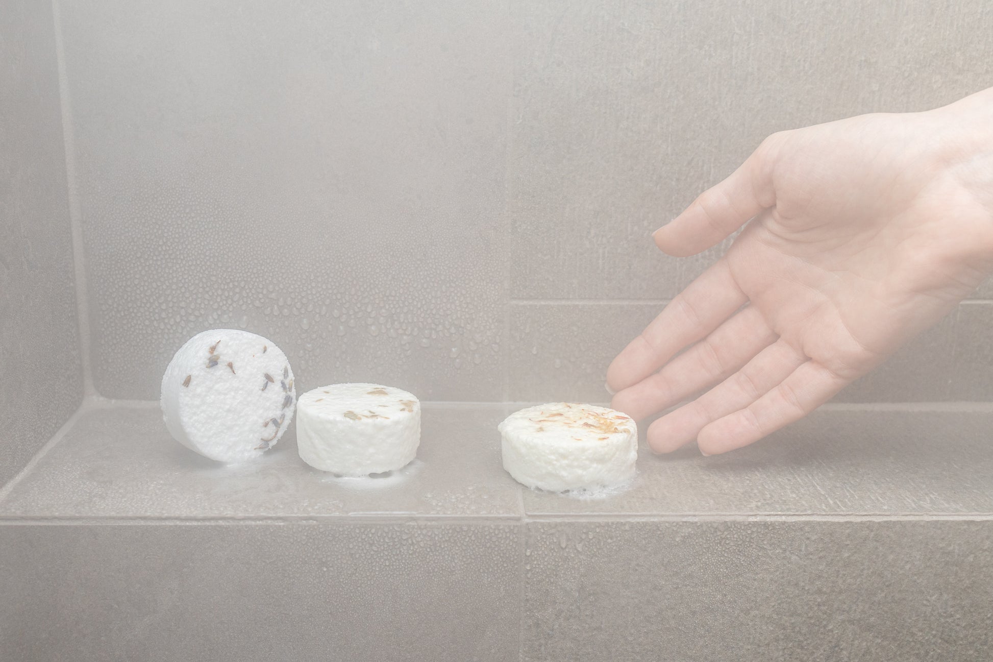 three shower steamers dissolving in the shower with steam and a hand reaching for one of the steamers