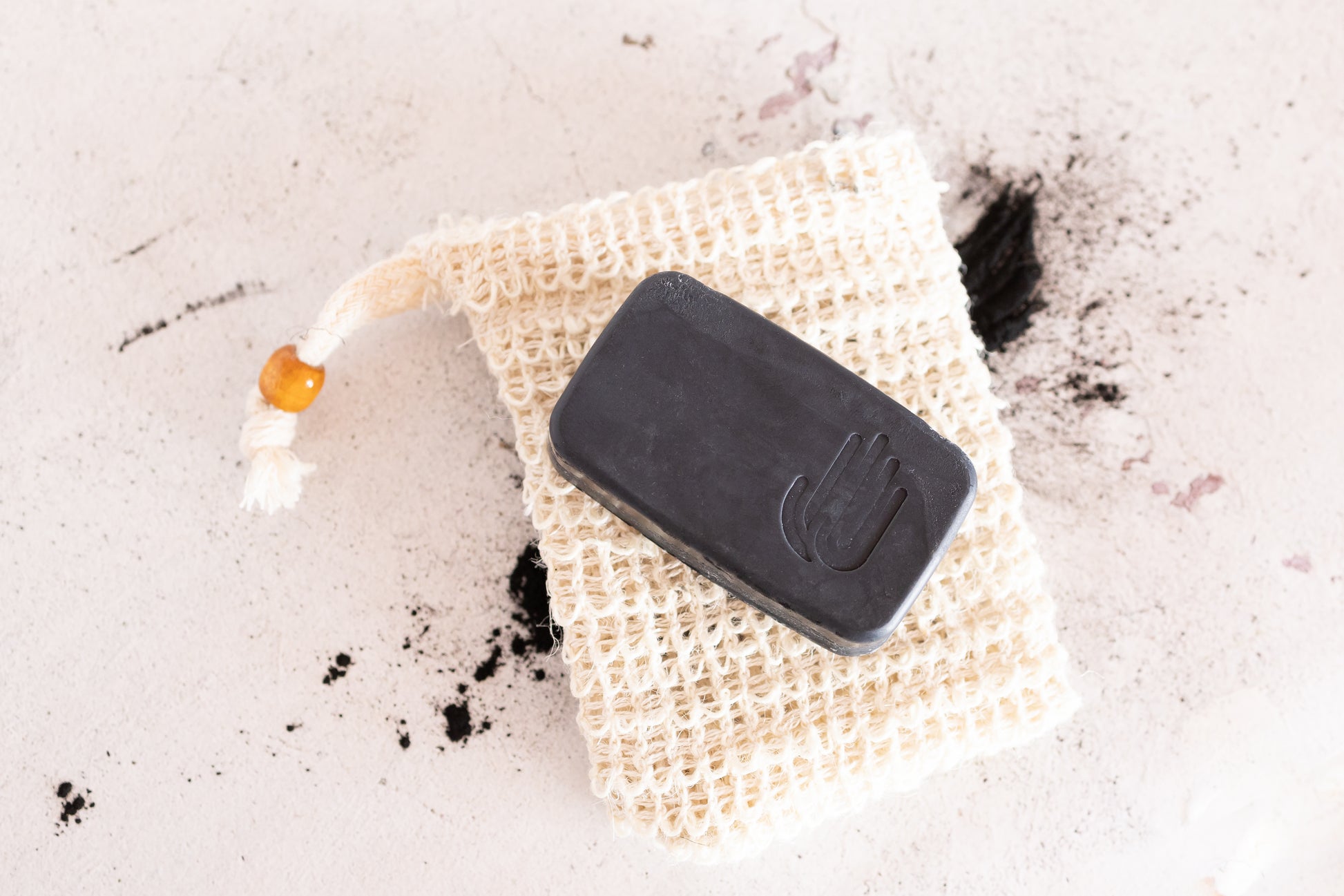 natural sisal soap saver bag with a wooden bead on the drawstring and a black soap sitting on it and charcoal scattered