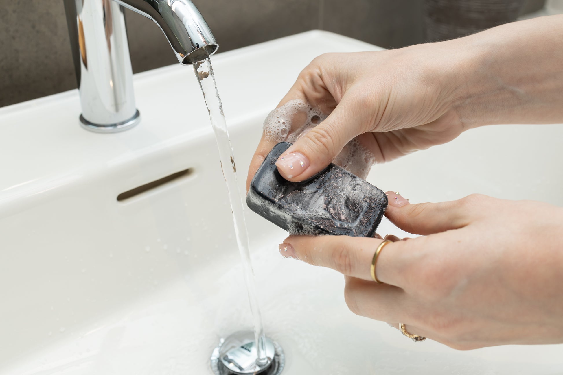 black tea tree charcoal square soap with suds being held by two hands under running water in a bathroom sink