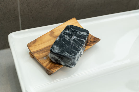 black tea tree charcoal square soap with suds sitting on a wooden soap dish on the corner of a bathroom sink