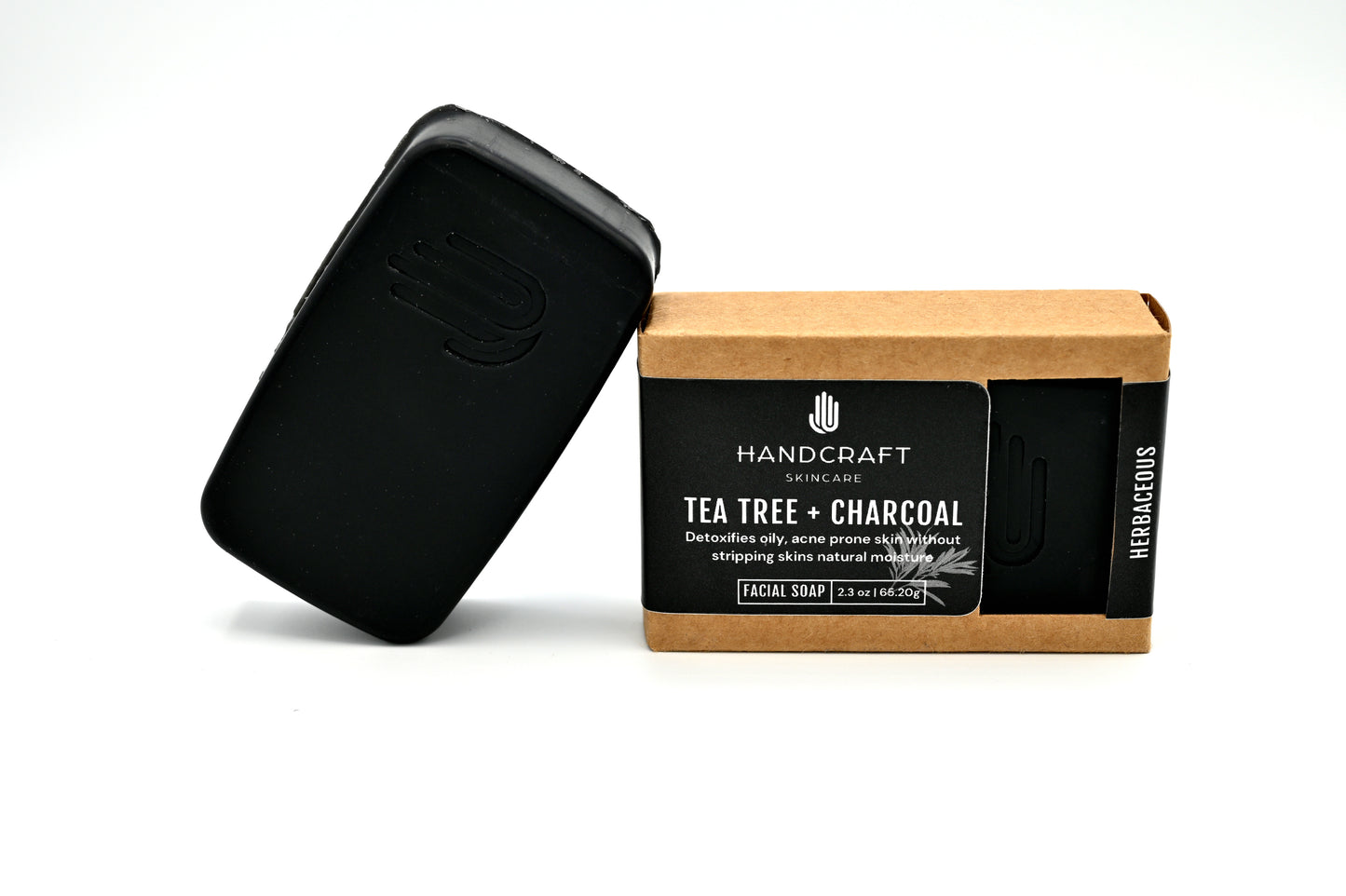 black tea tree and charcoal soap is unpackaged and leaning at an angle on a packaged tea tree and charcoal soap in a Kraft box with a black label